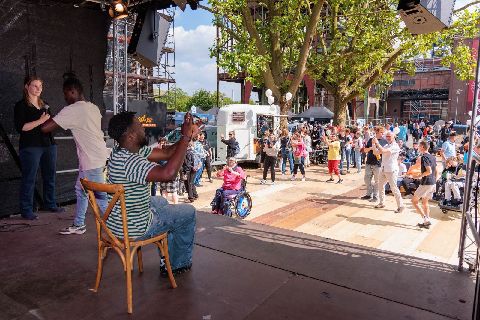 Dodzi Dougban leads a dance workshop on the outdoor stage at Zeche Ewald, accompanied by a sign language interpreter and dancing participants.