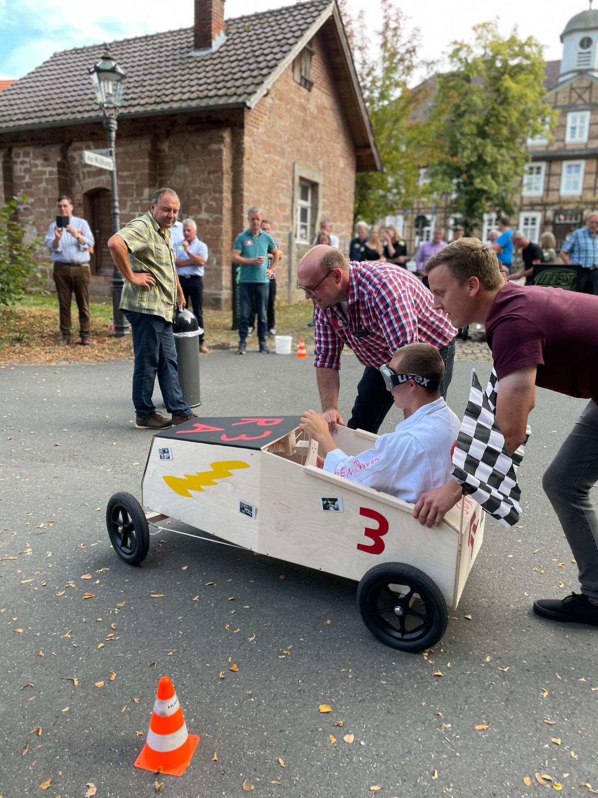 Teilnehmer eines Teambuilding-Events schieben eine selbstgebaute Seifenkiste &uuml;ber einen Parcours, w&auml;hrend weitere Personen die Aktion verfolgen.