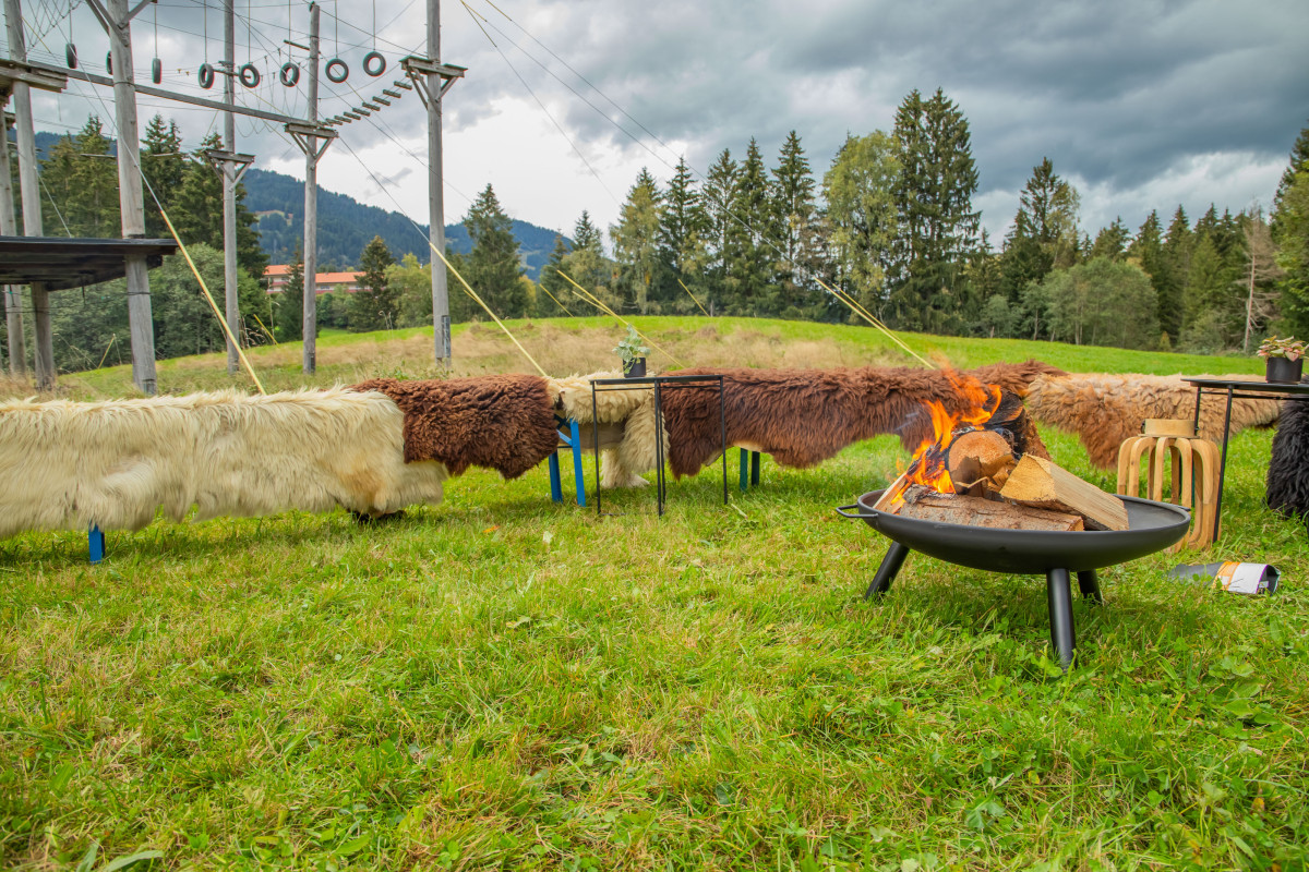 Loungebereich mit Sitzm&ouml;beln und Schafsfellen rund um eine brennende Feuerschale auf der Wiese der Pop-up-Location Bolsterlang.