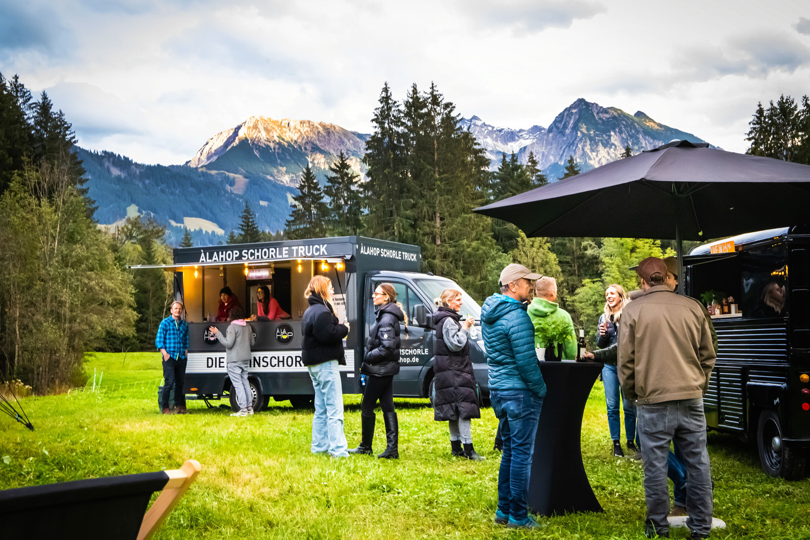 G&auml;ste stehen an zwei schwarzen Foodtrucks auf der PopUp Location Bolsterlang vor der Bergkulisse der Allg&auml;uer Alpen.