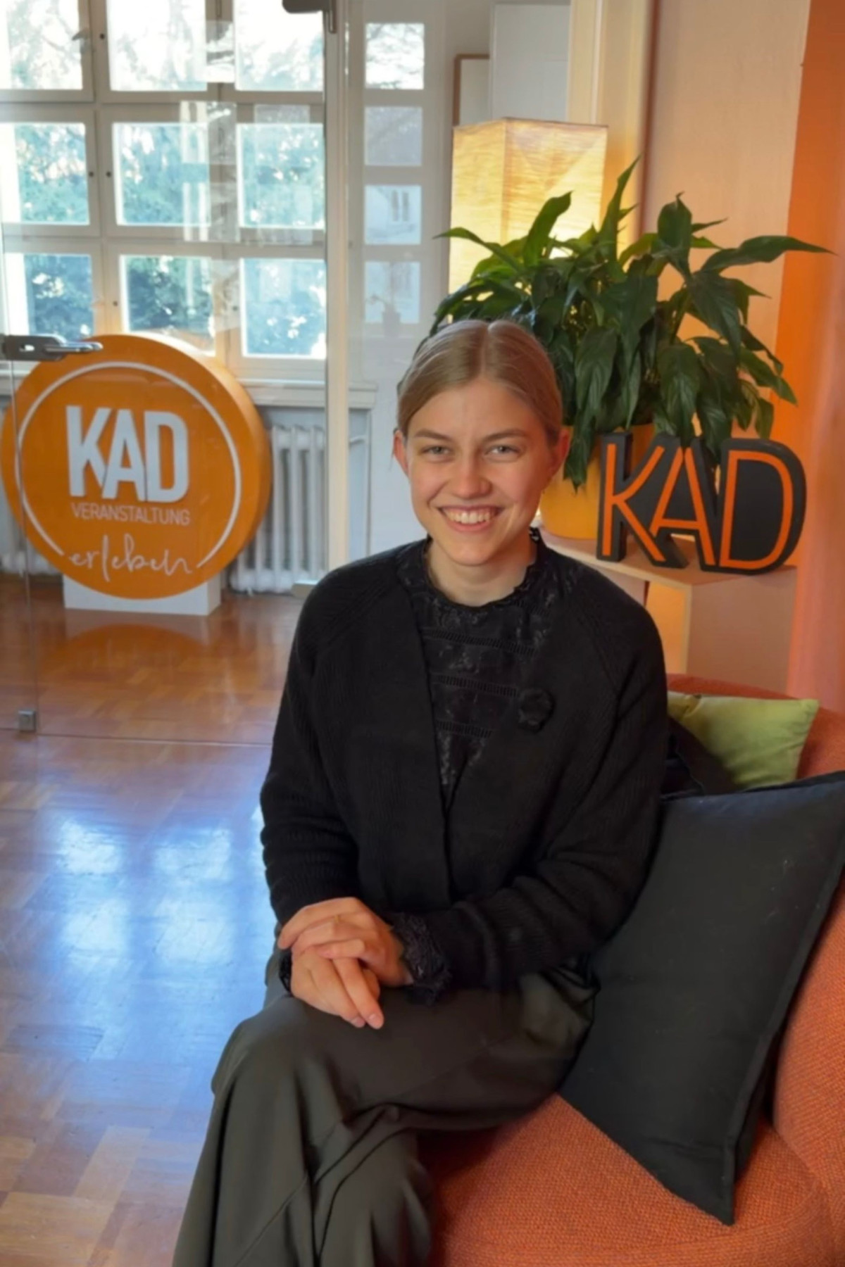 Dual student Veronika smiles while sitting on a sofa at the KAD office in Kempten, with orange KAD logos in the background.