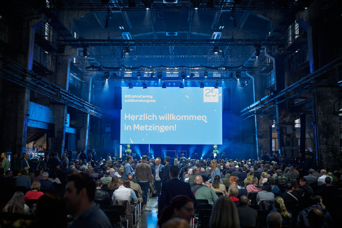 Blick in die Eventhalle mit beleuchteter B&uuml;hne und versammelten G&auml;sten.