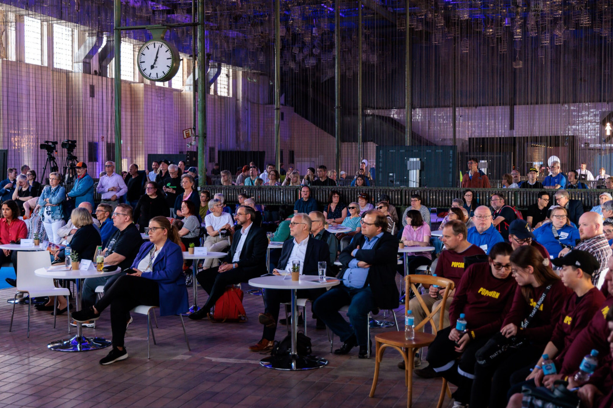 Audience sits attentively in front of the stage inside the industrial hall.