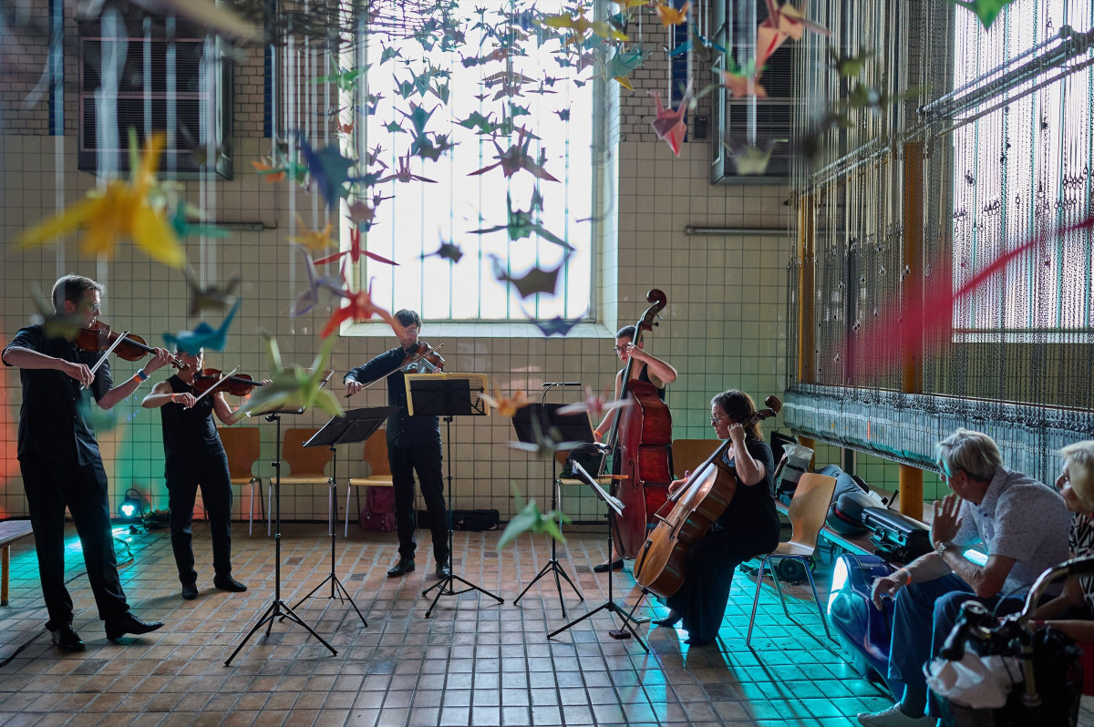 Chamber music performance in an industrial hall with hanging paper cranes and an attentive audience.