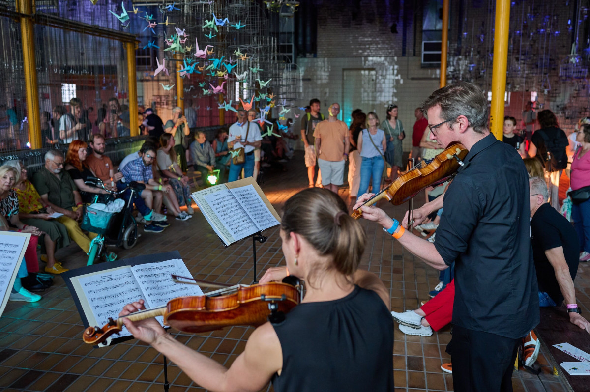 Chamber music performance in an industrial hall with hanging paper cranes and an attentive audience.